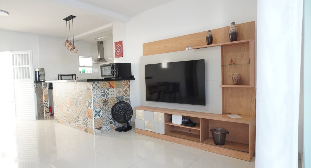 Sala de estar moderna de casa de praia em Juquehy com painel de TV em madeira, TV de tela plana, ventilador e bancada de cozinha integrada.