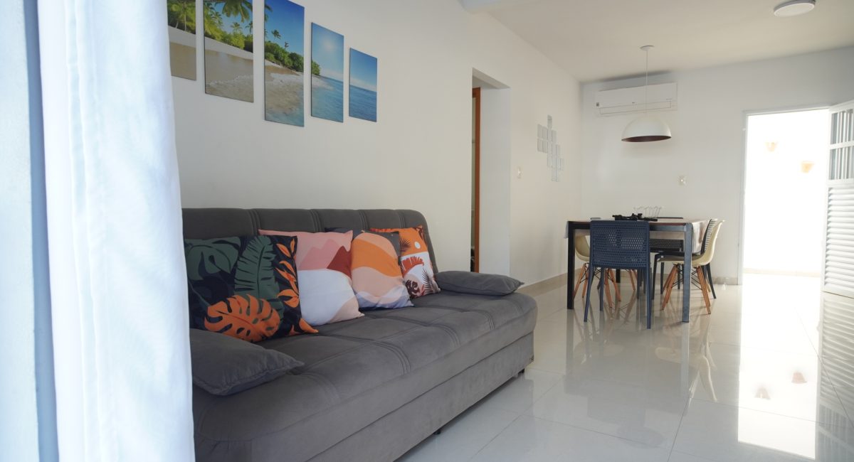 Sofá cinza confortável na sala de estar da casa de veraneio em Juquehy com almofadas decorativas e quadros de praia na parede branca.